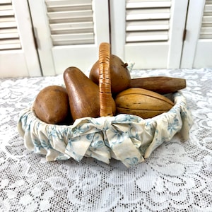 May include: A woven basket with a handle, filled with wooden fruit sculptures. The basket is lined with a fabric featuring a blue floral pattern. The wooden fruit includes pears, apples, and other shapes.