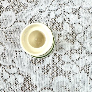 May include: A small, off-white ceramic egg cup with a green trim. The egg cup is set against a white lace tablecloth.