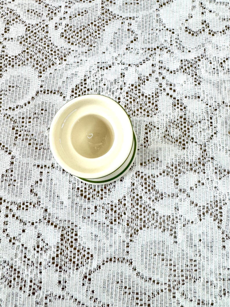 May include: A small, off-white ceramic egg cup with a green trim. The egg cup is set against a white lace tablecloth.