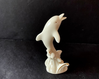 May include: A white porcelain dolphin figurine with gold accents, leaping out of the water. The dolphin is positioned on a base with a wave design.