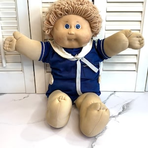 May include: A vintage Cabbage Patch Kid doll with blonde yarn hair, blue eyes, and a blue sailor outfit. The doll is sitting with its arms outstretched.