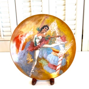 May include: A porcelain plate with a colorful painting of a ballerina in a white tutu and pink feathered skirt. The ballerina is dancing on a stage with colorful lights and a candy cane. The plate is signed "Emile Zolnier" in the lower right corner.