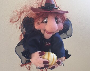 May include: A handmade witch doll with red hair, a black and red witch hat, and a blue dress. She is holding a small, yellow pumpkin.