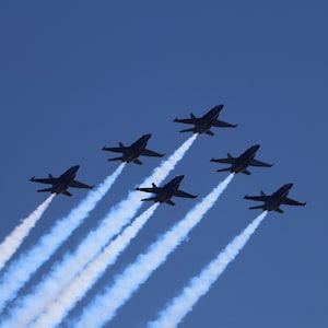May include: Six blue fighter jets fly in formation, leaving white smoke trails against a clear blue sky.