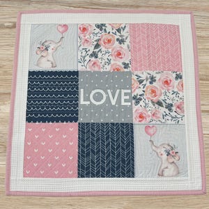 May include: A square patchwork quilt featuring nine panels with pink, gray, and navy blue colors. The design includes floral patterns, chevron stripes, heart motifs, and two illustrations of elephants holding heart-shaped balloons with the word LOVE.