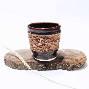 May include: A small, brown ceramic cup with a textured, crackled glaze. The cup is sitting on a wooden surface.