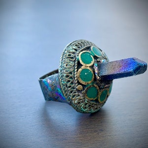 Könnte beinhalten: Ein Ring mit einem blauen und schillernden Stein, der in einem goldfarbenen Band mit grünen Emaille-Akzenten gefasst ist.