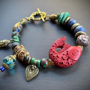 Könnte beinhalten: Armband mit einer roten, geschnitzten Fisch-Charm, goldfarbenen Akzenten und blauen, grünen und braunen Perlen. Enthält einen Buddha-Anhänger und einen Knebelverschluss. Ca. 20 cm lang.