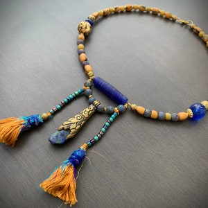 Op de afbeelding: Een meerstrengs ketting met blauwe, gouden en bruine kralen. De ketting heeft een lange, blauwe glazen kraal en twee kwastjes met gouden en blauwe accenten.
