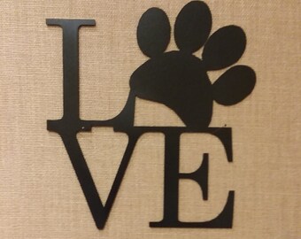 May include: Black metal sign with the word "LOVE" and a paw print replacing the "O". The sign is mounted on a tan, textured background. A decorative item for pet lovers.