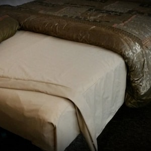 May include: A bed with a beige fitted sheet and a patterned comforter in shades of brown and gold. A green pillow with a brown border rests on the bed. The bed is in a dimly lit room.