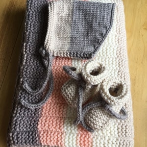 May include: A knitted baby set in shades of grey, pink, and white. The set includes a blanket, bonnet, and booties. The blanket is folded, with the bonnet and booties placed on top. The booties have grey ties.