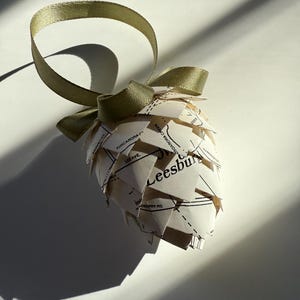 May include: A handmade ornament shaped like a pinecone, crafted from map paper. The ornament features the word "Leesburg" and other map details. It is adorned with a green ribbon and bow for hanging.