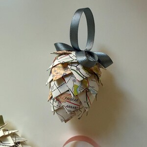 May include: A handmade Christmas ornament shaped like a pinecone. The ornament is constructed from map paper and has a gray ribbon for hanging. The map paper shows street names and numbers. The ornament is a unique holiday decoration.