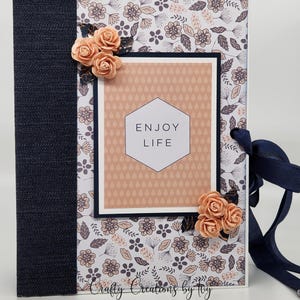May include: A small, peach-colored journal with a floral pattern and a navy blue ribbon. The front cover has a peach-colored frame with the text "ENJOY LIFE" in a white hexagon.
