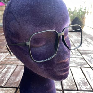 Könnte beinhalten: Ein Mannequin-Kopf mit schwarzen, quadratischen Brillen und einer violetten Samt-Bedeckung.