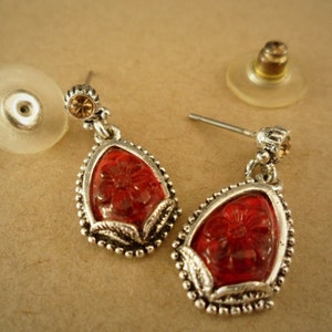 May include: A pair of silver dangle earrings with red glass teardrop-shaped stones. The stones have a floral design and are accented with small clear rhinestones.