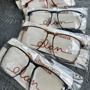 May include: Several pairs of eyeglasses in plastic packaging. The frames are in various colors, including black, brown, and burgundy. Each pair has a rectangular shape and is labeled with "The Elan Collection".