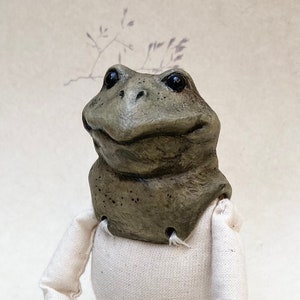 Op de afbeelding: Een handgemaakte kikkerpop met een grijze kop en ledematen, gekleed in een witte outfit. De kikker zit met uitgestrekte benen. De achtergrond is lichtbeige met een bloemenpatroon.