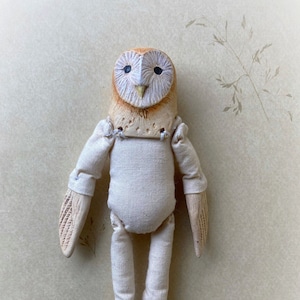Puede incluir: Una muñeca de búho de granero hecha a mano con un cuerpo de tela blanca y detalles pintados en marrón. El búho tiene una cara blanca con ojos negros y un pico marrón. La muñeca está posada con las alas extendidas y las piernas dobladas.
