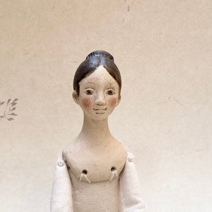 May include: A handcrafted doll with a calm expression, dressed in a white outfit and black socks. The doll features a detailed face with rosy cheeks and dark hair. The doll is seated with its legs outstretched.