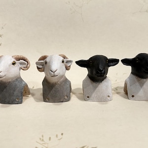 Puede incluir: Cuatro figuras de ovejas hechas a mano. Dos tienen caras blancas y cuernos rizados, con cuerpos grises y blancos. Las otras dos tienen caras y orejas negras, con cuerpos blancos. Cada una tiene un pequeño agujero redondo en la base.