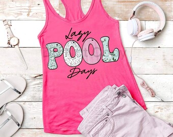 May include: Pink tank top with the text "Lazy Pool Days" in a floral font. The tank top is on a white wood background with a pair of silver sandals, a watch, and a pair of pink headphones.