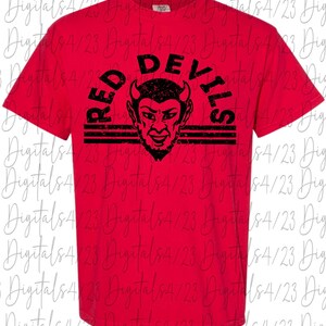 May include: A red t-shirt with the words "RED DEVILS" arched above a black devil face graphic. The graphic includes horns, a beard, and horizontal stripes. The shirt is a short-sleeved crew neck.