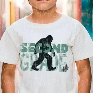 May include: White t-shirt with a black Bigfoot silhouette and the words "SECOND GRADE" in green block letters. Two small green trees are to the right of the word "GRADE".