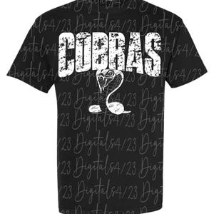 Puede incluir: Camiseta negra con la palabra "COBRAS" en letras blancas desgastadas y un gráfico de cobra blanca. La camiseta tiene cuello redondo y mangas cortas. El diseño está centrado.