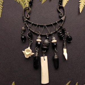 May include: A handcrafted necklace with a dark metal wire frame and chain. The necklace features black beads, bone, and a tooth pendant. The design includes a variety of shapes and textures, creating a unique, gothic-inspired accessory.