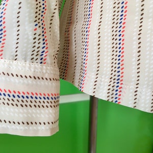 Op de afbeelding: Een wit overhemd met lange mouwen met een bruin, rood en blauw gestreept patroon. Het shirt heeft een gestippeld patroon binnen de strepen.