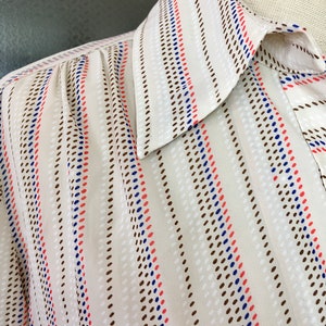 Op de afbeelding: Een wit shirt met een gestreept patroon met bruine, blauwe en rode stippen. Het shirt heeft een puntige kraag.