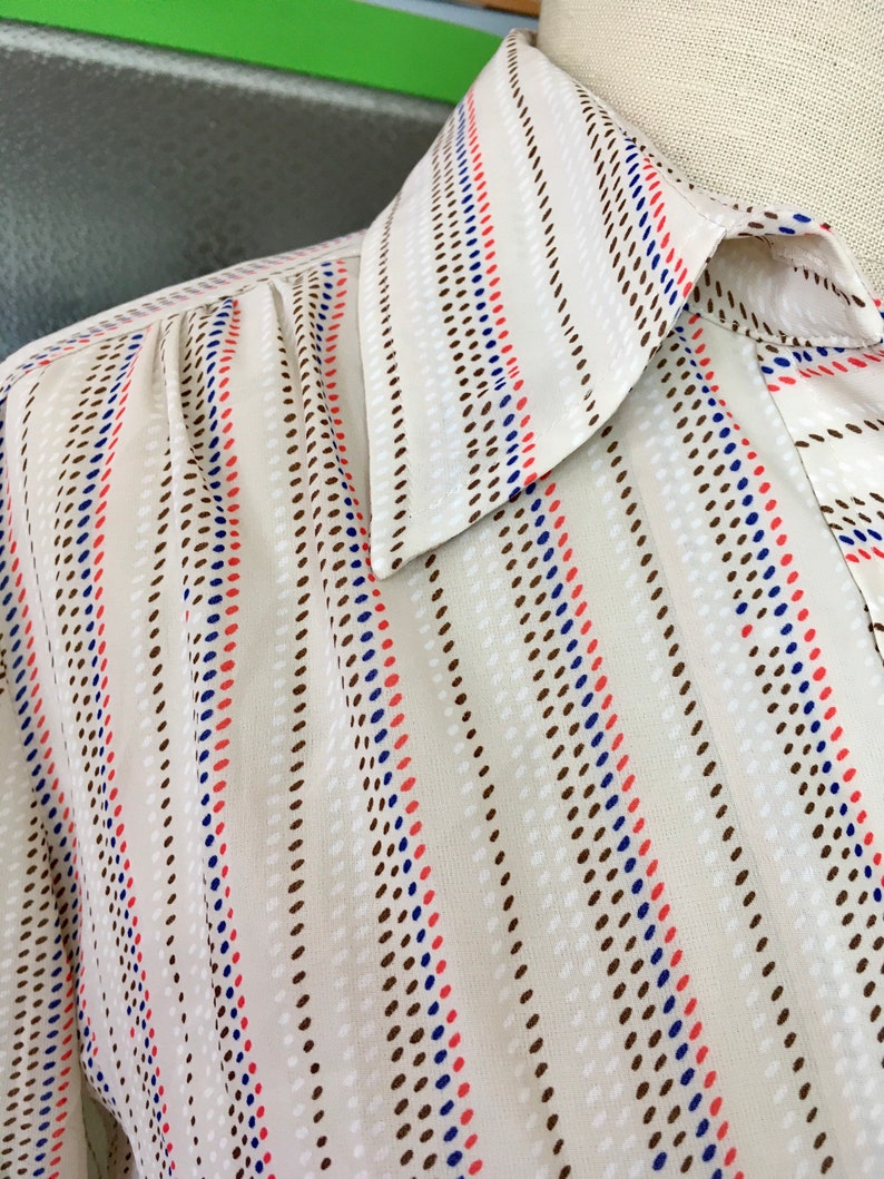 Op de afbeelding: Een wit shirt met een gestreept patroon met bruine, blauwe en rode stippen. Het shirt heeft een puntige kraag.