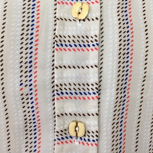 Op de afbeelding: Close-up van een beige overhemd met knopen met bruine, blauwe en rode strepen. Het overhemd heeft twee gouden knopen.