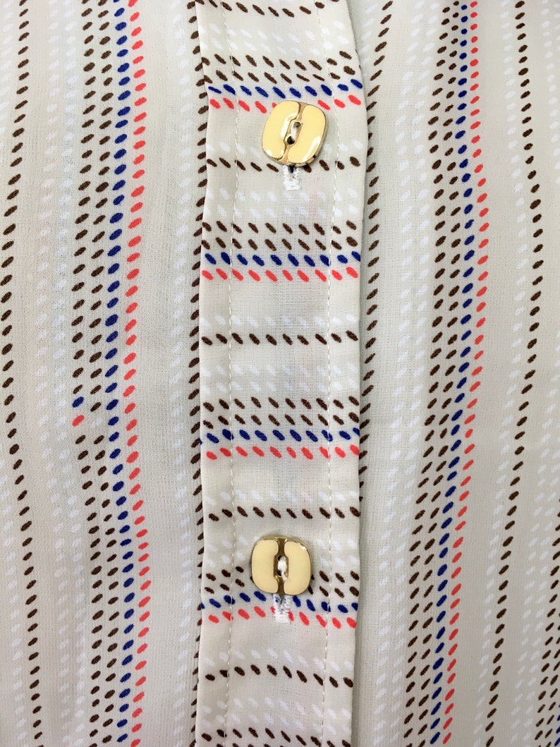 Op de afbeelding: Close-up van een beige overhemd met knopen met bruine, blauwe en rode strepen. Het overhemd heeft twee gouden knopen.