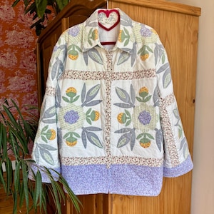 May include: A quilted jacket with a floral pattern in white, green, purple, and yellow. The jacket has a button-up front and a collar. The floral design includes leaves and flower shapes. The jacket is hanging on a red heart-shaped hanger.