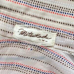 Op de afbeelding: Een witte stof met een bruin, blauw en rood stippenpatroon. De stof heeft een wit label met de tekst "Mitsukoshi - M" erop.