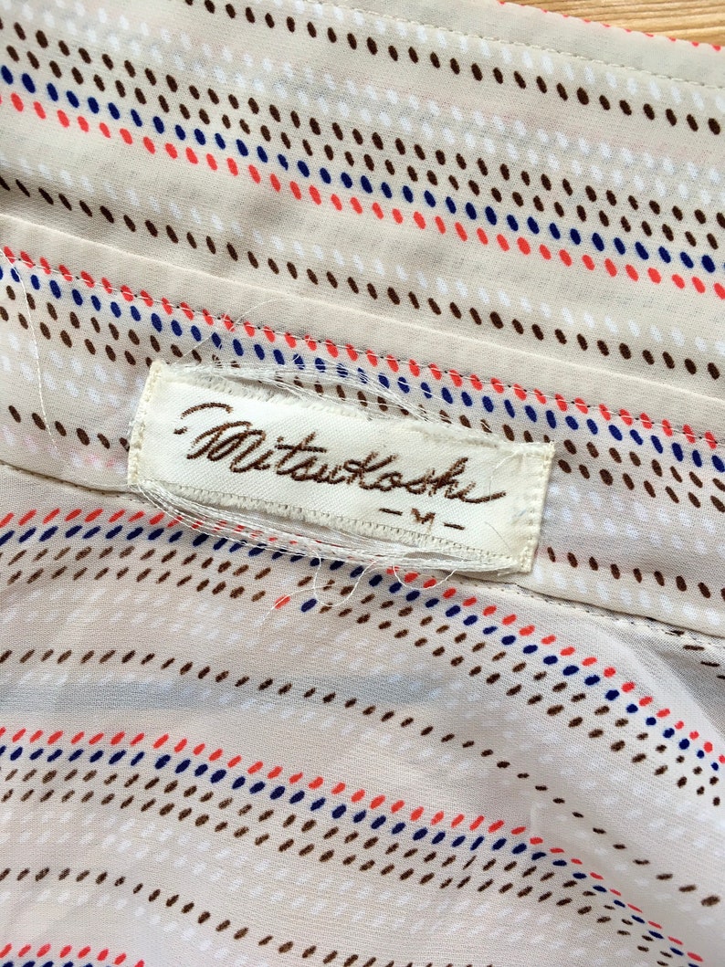 Op de afbeelding: Een witte stof met een bruin, blauw en rood stippenpatroon. De stof heeft een wit label met de tekst "Mitsukoshi - M" erop.