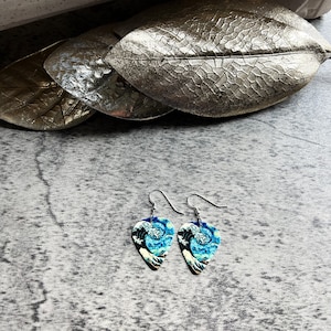 K&ouml;nnte beinhalten: Ein Paar silberfarbene Ohrh&auml;nger. Jeder Ohrring hat die Form eines Gitarrenplektrums und zeigt ein blau-wei&szlig;es Wellenmuster. Die Ohrringe werden auf einer strukturierten grauen Oberfl&auml;che pr&auml;sentiert, mit silberfarbenen blattf&ouml;rmigen Dekorationen im Hintergrund.