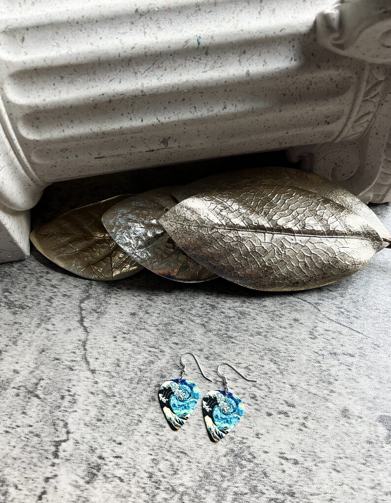 K&ouml;nnte beinhalten: Ein Paar Ohrringe mit einem blau-wei&szlig;en Wellenmuster. Die Ohrringe sind wie Plektren geformt und haben silberfarbene Haken. Die Ohrringe werden auf einer grauen Oberfl&auml;che pr&auml;sentiert.