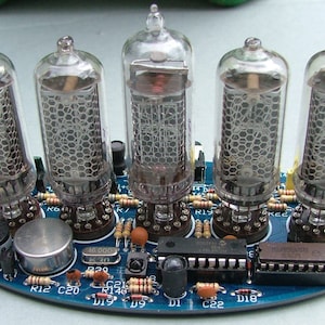 Pode incluir: Uma placa de circuito azul com cinco tubos Nixie de vidro transparente, um pequeno alto-falante e alguns circuitos integrados. A placa tem vários resistores e capacitores etiquetados com números e letras.