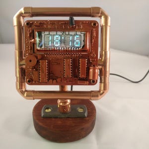 Puede incluir: Un reloj digital con una pantalla de circuito impreso rectangular con números azules brillantes, encerrado en un marco cuadrado de tubería de cobre. El reloj está montado sobre una base de madera oscura con un conector metálico y un cable negro.