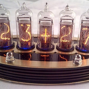 Könnte beinhalten: Eine Digitaluhr mit fünf Glasröhren, die beleuchtete Zahlen anzeigen. Die Uhr hat eine Messing- und schwarze Basis mit sichtbarer Verkabelung und Metallbeschlägen. Die Zahlen leuchten orange.