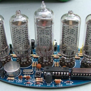 May include: A blue circuit board with five clear glass vacuum tubes. The tubes have a honeycomb-like internal structure. Various electronic components, including resistors and chips, are visible on the board. The board has a small speaker and a metal disc.