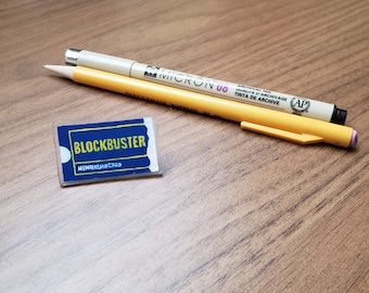 May include: A yellow pencil and a beige pen with black ink on a wooden surface. The pen has the words "Micron 00" and "Archival Ink" printed on it. A blue and white plastic card with the word "Blockbuster" and "Membership Card" printed on it is also on the surface.
