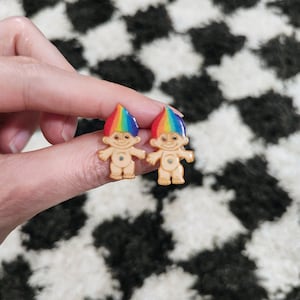 May include: A pair of rainbow troll earrings. The trolls are made of plastic and have a light brown skin tone. They are wearing rainbow colored hair and have blue eyes.