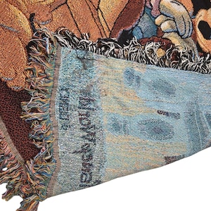May include: A woven throw blanket featuring a colorful graphic of Mickey Mouse and other characters. The fabric has a textured, tapestry-style weave with visible fringe edges. The reverse side shows a lighter, muted blue tone with the text Snow White.