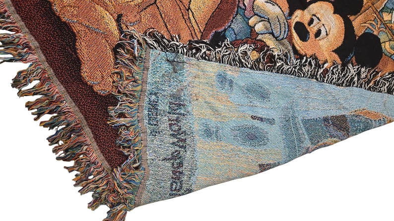 May include: A woven throw blanket featuring a colorful graphic of Mickey Mouse and other characters. The fabric has a textured, tapestry-style weave with visible fringe edges. The reverse side shows a lighter, muted blue tone with the text Snow White.