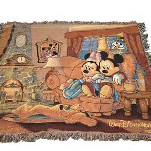 May include: A woven tapestry throw blanket featuring a scene of Mickey and Minnie Mouse sitting on a sofa by a stone fireplace with Pluto sleeping on the floor. The design includes a window with a snowman outside and the text Walt Disney World.